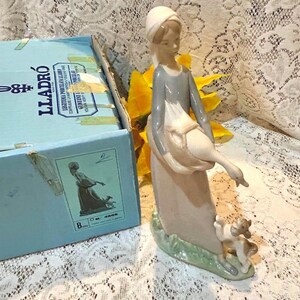 May include: A porcelain figurine of a woman in a long dress and head covering, holding a goose and accompanied by two small dogs. The figurine is on a green base. A blue box with the word "LLADRÓ" is in the background.
