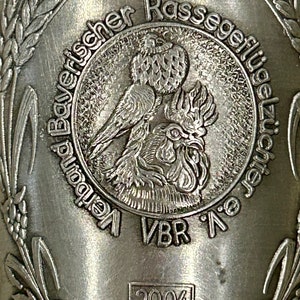 Op de afbeelding: Close-up van een zilverkleurig metalen object met reli&euml;ftekst en afbeeldingen. Het centrale ontwerp toont een vogel en een haan. De tekst bevat "Bayerischer Rassegefl&uuml;gelz&uuml;chter" en "VBR". Het jaar "2004" is ook zichtbaar.