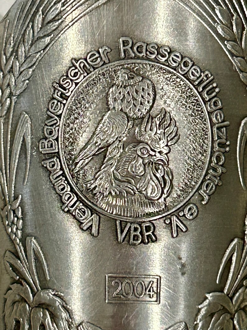 Op de afbeelding: Close-up van een zilverkleurig metalen object met reli&euml;ftekst en afbeeldingen. Het centrale ontwerp toont een vogel en een haan. De tekst bevat "Bayerischer Rassegefl&uuml;gelz&uuml;chter" en "VBR". Het jaar "2004" is ook zichtbaar.