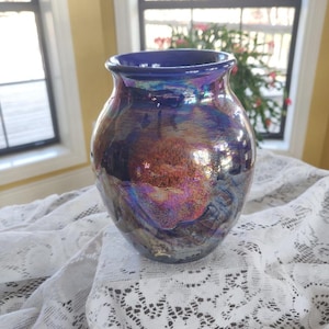 May include: A handcrafted ceramic vase with a glossy, iridescent finish. The vase features a dark blue rim and a body with shades of purple, bronze, and silver. The vase is sitting on a white lace tablecloth.