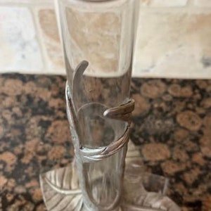 May include: A clear glass bud vase with a silver-colored metal leaf design base and accents. The vase is cylindrical and stands upright. The base features three stylized leaves. The vase is approximately 8 inches tall.