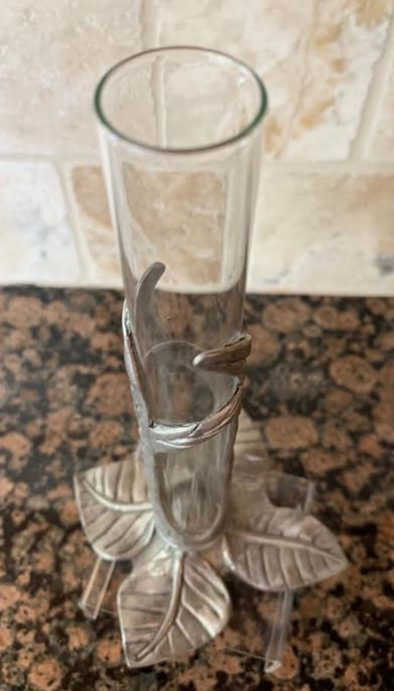 May include: A clear glass bud vase with a silver-colored metal leaf design base and accents. The vase is cylindrical and stands upright. The base features three stylized leaves. The vase is approximately 8 inches tall.