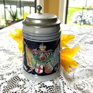 May include: A ceramic beer stein with a dark blue base, adorned with heraldic crests and a silver-colored metal lid. The stein features intricate detailing and a decorative handle. The background includes a white lace tablecloth and yellow flowers.