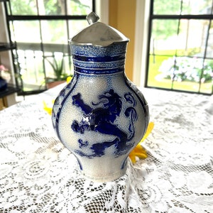 May include: A blue and white ceramic pitcher with a pewter lid. The pitcher features a raised lion design and blue geometric patterns. The pitcher is sitting on a white lace tablecloth.