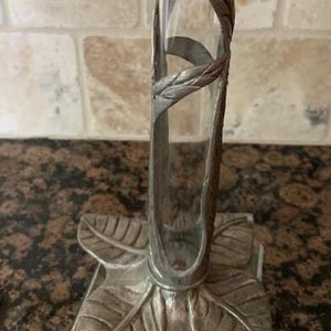 May include: A silver-colored bud vase with a clear glass tube. The vase has a decorative metal frame with a leaf-shaped base and a curved design around the glass. The vase is designed to hold a single flower or small floral arrangement.