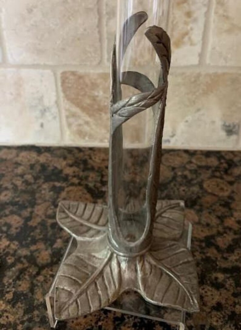 May include: A silver-colored bud vase with a clear glass tube. The vase has a decorative metal frame with a leaf-shaped base and a curved design around the glass. The vase is designed to hold a single flower or small floral arrangement.