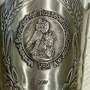 Op de afbeelding: Een zilverkleurig metalen drinkglas met een reli&euml;f ontwerp. Het ontwerp bevat de tekst "Bayerischer Rassegefl&uuml;gelz&uuml;chter e.V." en het jaar "2007". Het glas toont afbeeldingen van tarwe en een haan.