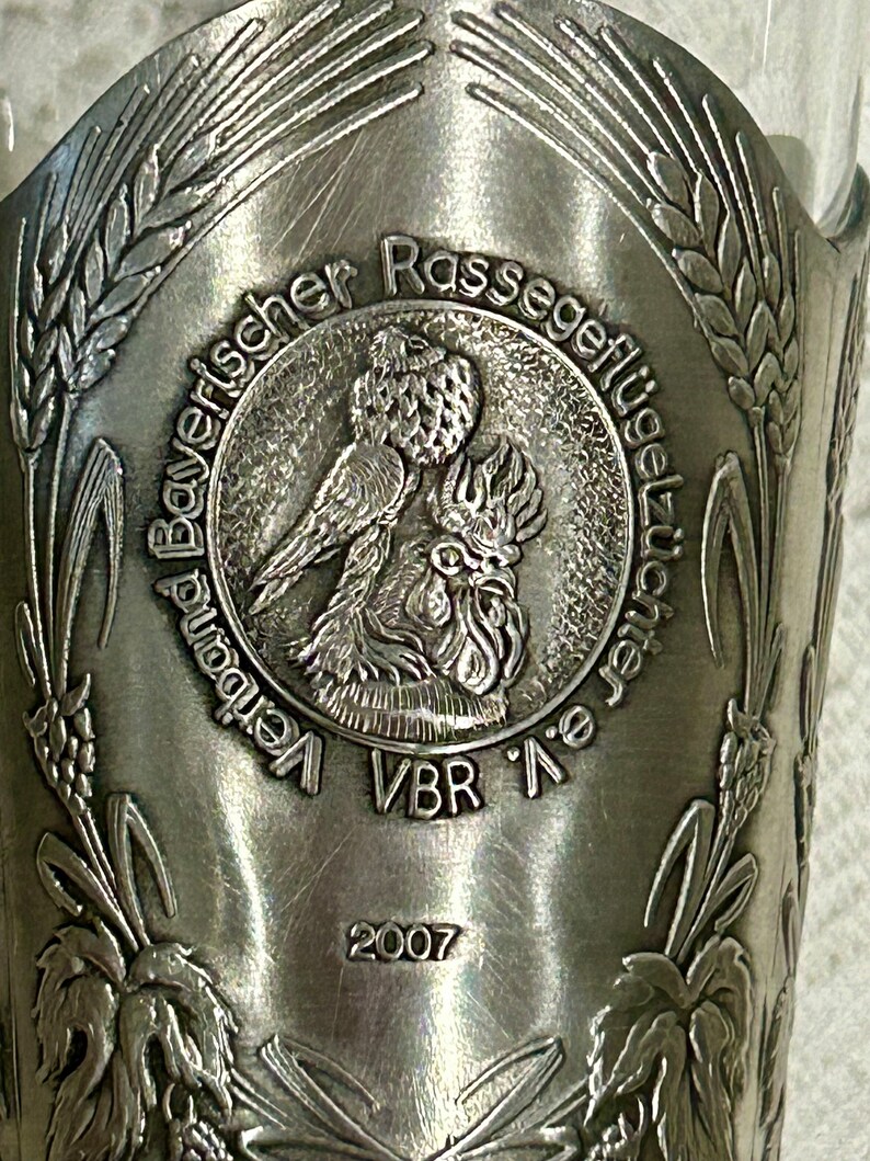 Op de afbeelding: Een zilverkleurig metalen drinkglas met een reli&euml;f ontwerp. Het ontwerp bevat de tekst "Bayerischer Rassegefl&uuml;gelz&uuml;chter e.V." en het jaar "2007". Het glas toont afbeeldingen van tarwe en een haan.