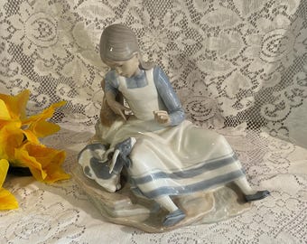 May include: A porcelain figurine of a girl seated on a rock, wearing a white dress with blue accents and a blue long-sleeved shirt. The girl is interacting with a small, gray and white animal. The figurine is a decorative item.