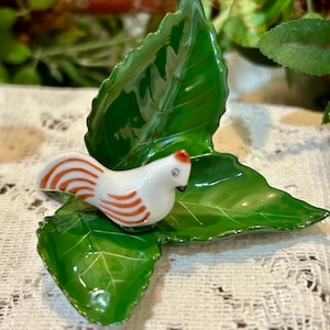 May include: A ceramic figurine of a small white bird with orange stripes and a red crest, perched on a glossy green leaf. The leaf is part of a larger, stylized leaf arrangement, creating a nature-inspired decorative piece.