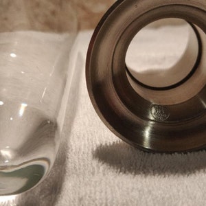 Op de afbeelding: Een heldere glazen vaas en een zilveren metalen kandelaar worden getoond. De vaas is cilindrisch met een afgeronde bodem. De kandelaar heeft een cirkelvormige opening en een gedetailleerde basis. De items staan op een witte ondergrond.