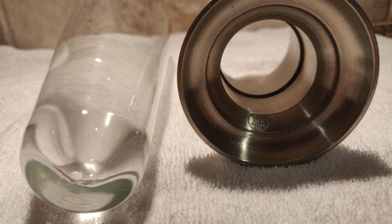 Op de afbeelding: Een heldere glazen vaas en een zilveren metalen kandelaar worden getoond. De vaas is cilindrisch met een afgeronde bodem. De kandelaar heeft een cirkelvormige opening en een gedetailleerde basis. De items staan op een witte ondergrond.