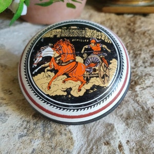 May include: A round, decorative container with a black, red, and gold design. The lid features a chariot scene with the word "ACHILLES". The horses are orange and white, and the background is black and gold.