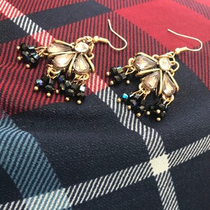 Pode incluir: Um par de brincos de ouro com um design de flor de cristal transparente e contas pretas penduradas na parte inferior.