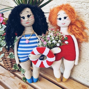 May include: Two crocheted dolls wearing swimsuits and holding a life preserver and a bouquet of flowers. The doll on the left has black curly hair and is wearing a blue and white striped swimsuit. The doll on the right has red curly hair and is wearing a red swimsuit.