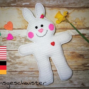 May include: White crocheted bunny rabbit toy with pink cheeks, a pink nose, and a pink flower on its head. The bunny is sitting on a wooden surface with a yellow flower and hearts.