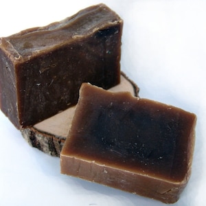 Può includere: Due saponette marroni su una fetta di legno. Il sapone è di colore marrone scuro e ha una consistenza ruvida.