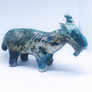 Op de afbeelding: Een keramieken geitenbeeldje met een blauw-witte glazuur. De sculptuur heeft een getextureerd oppervlak en staat op vier poten. De geit heeft hoorns en een open mond, wat het een unieke uitstraling geeft. Het beeldje is een decoratief item.