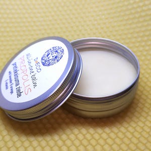 Puede incluir: Un bote plateado abierto con un bálsamo blanco cremoso. La tapa, parcialmente abierta, muestra texto como "DECO all-in-one balm PROPOLIS" y un diseño floral azul. Probablemente un producto para el cuidado de la piel.