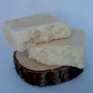 Puede incluir: Dos barras rectangulares de jabón artesanal, apiladas sobre una pequeña rodaja de madera redonda. El jabón es de color blanco crema con una superficie ligeramente texturizada. La rodaja de madera tiene un borde de corteza marrón oscuro.