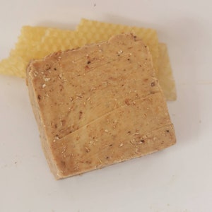 Könnte beinhalten: Ein rechteckiger Block handgemachter Seife in hellbrauner Farbe mit sichtbaren Flocken. Zwei Stücke gelbes Wabenwachs befinden sich hinter der Seife. Die Seife hat eine strukturierte Oberfläche.