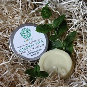 May include: A silver tin of natural deodorant with a white label that reads "DOECO natural deodorant GREEN TEA energy of nature with tea tree & clary sage essential oils". The tin is sitting on a bed of shredded paper with a sprig of green mint leaves.