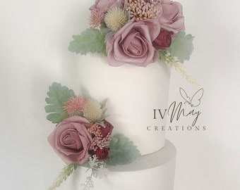 May include: A three-tiered white cake decorated with dusty rose and deep red artificial flowers, light green foliage, and spiky thistle accents. The text IV May CREATIONS with a butterfly icon is visible on the middle tier of the cake.