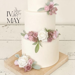 May include: A three-tiered white cake decorated with artificial dusty rose, mauve, and white foam roses, along with sage green leaves. The logo IV MAY CREATIONS is visible on the left. The cake is displayed on a light-coloured wooden board.