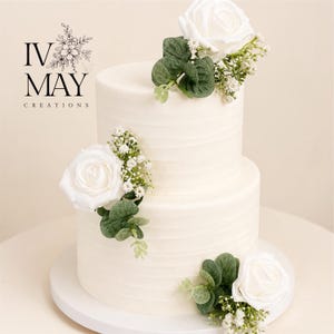 Op de afbeelding: Een witte taart met twee lagen en horizontaal getextureerd glazuur, versierd met witte rozen, groene eucalyptusbladeren en kleine witte gipskruidbloemen. Het logo IV MAY CREATIONS is zichtbaar in de linkerbovenhoek van de afbeelding.