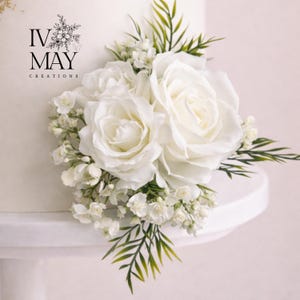 May include: A white cake decorated with a cluster of white roses, small white filler flowers, and green foliage. The text IV MAY CREATIONS with a floral graphic is printed on the side of the cake, which rests on a white cake stand.