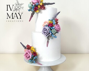 May include: A two-tier white cake decorated with clusters of artificial flowers in shades of pink, purple, blue, yellow, and green. The floral arrangements are placed on the top tier, middle section, and base. The text IV MAY CREATIONS is visible.