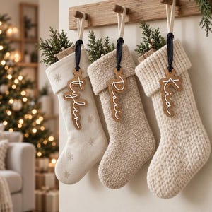 May include: Three cream-colored Christmas stockings hanging from a wooden rack. Each stocking features a wooden name tag tied with a black ribbon, displaying the names Tyler, Raxi, and Kia in white script. The stockings have varied textures.