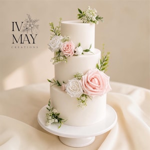 May include: A three-tiered white cake adorned with pink and white roses, greenery, and small white flowers. The cake is presented on a white cake stand. The logo "IV MAY CREATIONS" is visible in the upper left corner.