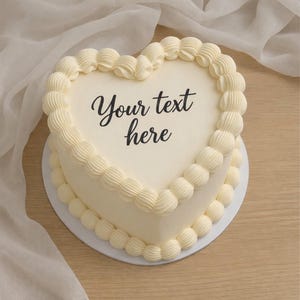 Op de afbeelding: Een hartvormige taart met gladde, gebroken witte glazuur, voorzien van decoratieve gespoten randen langs de boven- en onderkant. In het midden van de taart staat de zwarte tekst "Your text here" in een cursief lettertype.