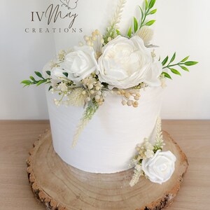 Op de afbeelding: Een witte taart met twee lagen, versierd met witte rozen, groen en gedroogde bloemen. De taart staat op een rustieke houten schijf. De tekst "IV May Creations" is zichtbaar.