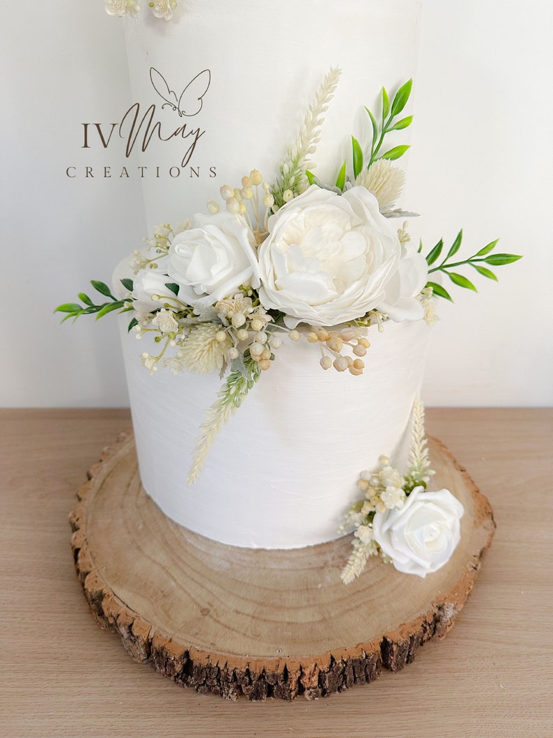 Op de afbeelding: Een witte taart met twee lagen, versierd met witte rozen, groen en gedroogde bloemen. De taart staat op een rustieke houten schijf. De tekst "IV May Creations" is zichtbaar.