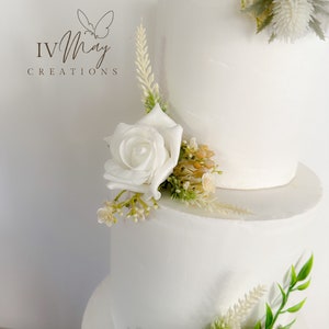 Op de afbeelding: Een witte taart met drie lagen, versierd met witte rozen en groen. De taart heeft een gladde afwerking en is versierd met diverse bloemstukken. Het logo "IV May Creations" is zichtbaar in de linkerbovenhoek.