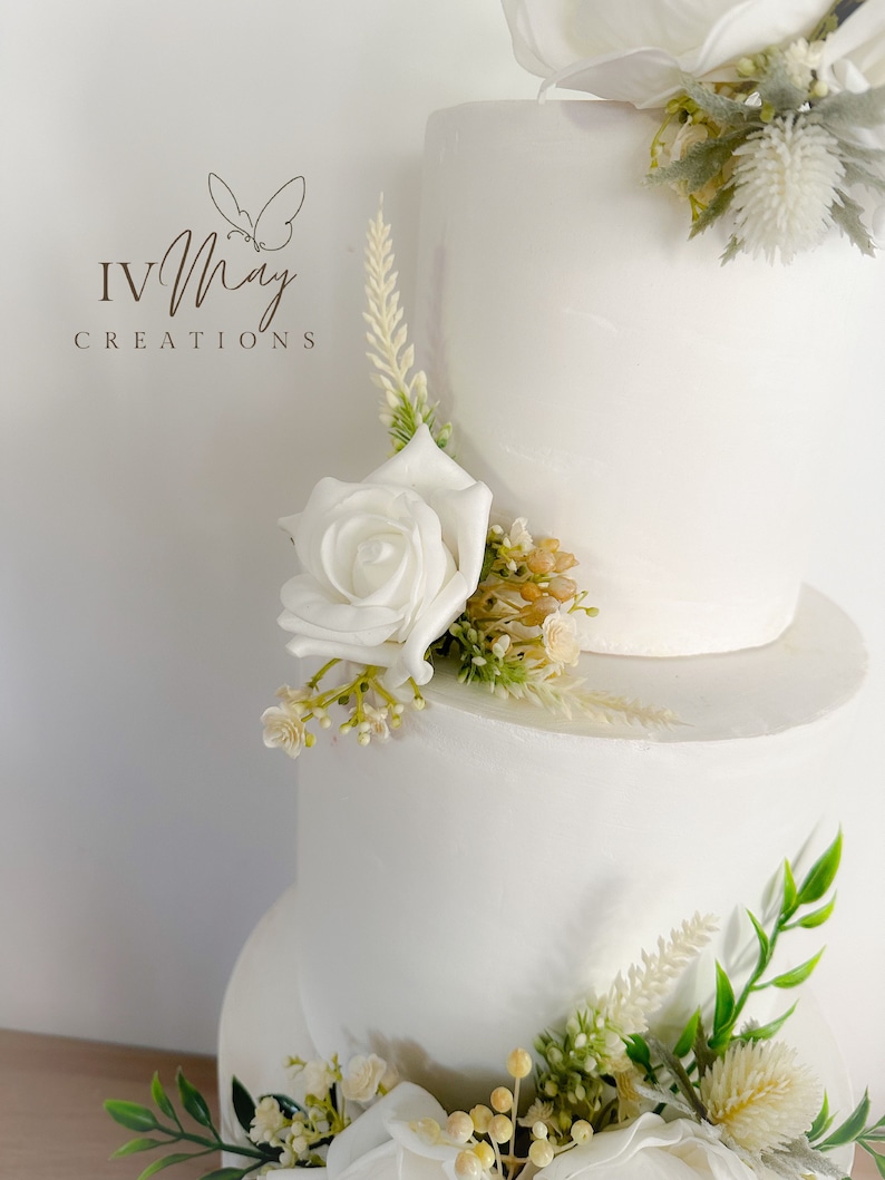 Op de afbeelding: Een witte taart met drie lagen, versierd met witte rozen en groen. De taart heeft een gladde afwerking en is versierd met diverse bloemstukken. Het logo "IV May Creations" is zichtbaar in de linkerbovenhoek.
