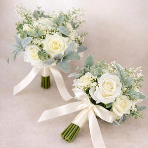 Op de afbeelding: Twee bloemboeketten met crèmekleurige rozen, kleine witte vulbloemen en saliegroene eucalyptusbladeren. Elk boeket is samengebonden met een glad, crèmekleurig satijnen lint dat strak om de stelen is gewikkeld voor een nette afwerking.