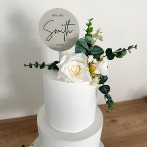 K&ouml;nnte beinhalten: Eine zweist&ouml;ckige wei&szlig;e Hochzeitstorte, verziert mit wei&szlig;en Rosen und Gr&uuml;n. Ein runder silberner Tortendecker mit dem Text "MR & MRS Smith 14.02.26" befindet sich oben auf der Torte.
