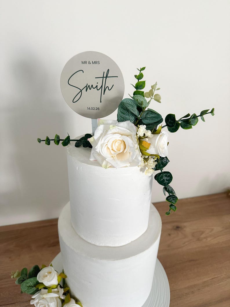 K&ouml;nnte beinhalten: Eine zweist&ouml;ckige wei&szlig;e Hochzeitstorte, verziert mit wei&szlig;en Rosen und Gr&uuml;n. Ein runder silberner Tortendecker mit dem Text "MR & MRS Smith 14.02.26" befindet sich oben auf der Torte.