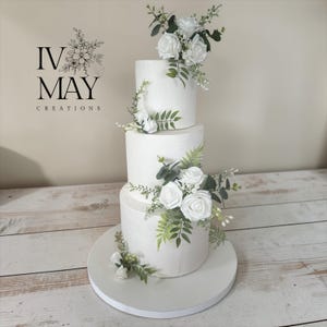 May include: A three-tier white cake decorated with white roses and green foliage. The text IV MAY CREATIONS is visible on the left. The cake is smooth with a subtle texture and sits on a round white base, featuring floral arrangements on each tier.