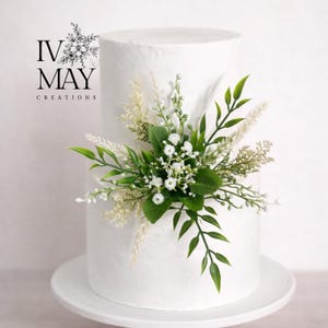 Op de afbeelding: Een witte taart met twee lagen, versierd met een centraal arrangement van kunstmatige groene bladeren, witte gipskruidbloemen en beige gevederde pluimen. De tekst IV MAY CREATIONS staat in het zwart linksboven in de afbeelding.