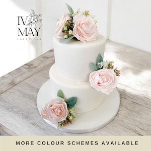 May include: A two-tier white cake decorated with light pink artificial roses, small white filler flowers, and green eucalyptus leaves. The text IV MAY CREATIONS appears in the upper left corner, and the text MORE COLOUR SCHEMES AVAILABLE is at the bottom.