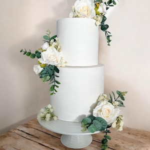 K&ouml;nnte beinhalten: Eine zweist&ouml;ckige wei&szlig;e Hochzeitstorte, verziert mit wei&szlig;en Rosen und Gr&uuml;n. Die Torte steht auf einem wei&szlig;en Tortenst&auml;nder und befindet sich auf einem Holztisch.