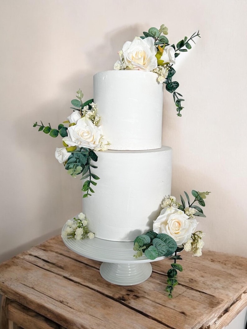 K&ouml;nnte beinhalten: Eine zweist&ouml;ckige wei&szlig;e Hochzeitstorte, verziert mit wei&szlig;en Rosen und Gr&uuml;n. Die Torte steht auf einem wei&szlig;en Tortenst&auml;nder und befindet sich auf einem Holztisch.