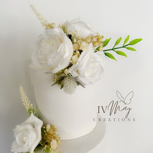 Op de afbeelding: Een witte taart met meerdere lagen, versierd met witte rozen, pioenrozen en groen. De taart heeft de tekst "IV May Creations" met een vlinderlogo. Een klassiek ontwerp voor feesten.