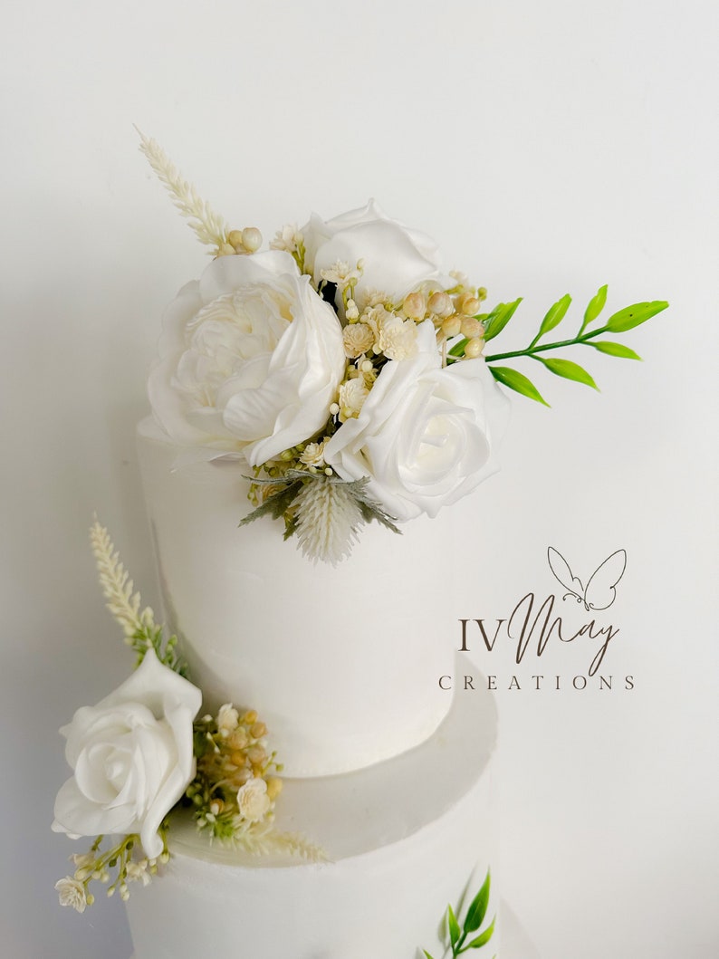 Op de afbeelding: Een witte taart met meerdere lagen, versierd met witte rozen, pioenrozen en groen. De taart heeft de tekst "IV May Creations" met een vlinderlogo. Een klassiek ontwerp voor feesten.