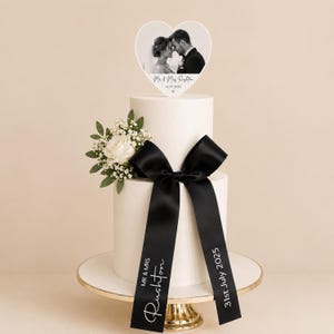 May include: A two-tier white cake featuring a black satin ribbon tied in a bow, adorned with a white rose and baby's breath. The ribbon displays the text "MR & MRS RUSHTON" and "31st July 2025". A heart-shaped topper shows a black and white photo.