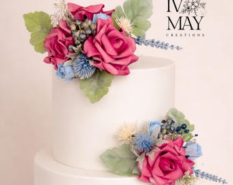 May include: A two-tiered white cake decorated with artificial floral arrangements featuring deep pink roses, light blue rosebuds, blue thistle, small dark berries, and pale green leaves. The text IV MAY CREATIONS is visible in the upper right corner.